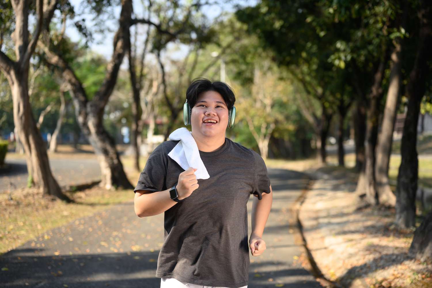 A smiling person wearing headphones and a towel jogging along a tree-lined park path.