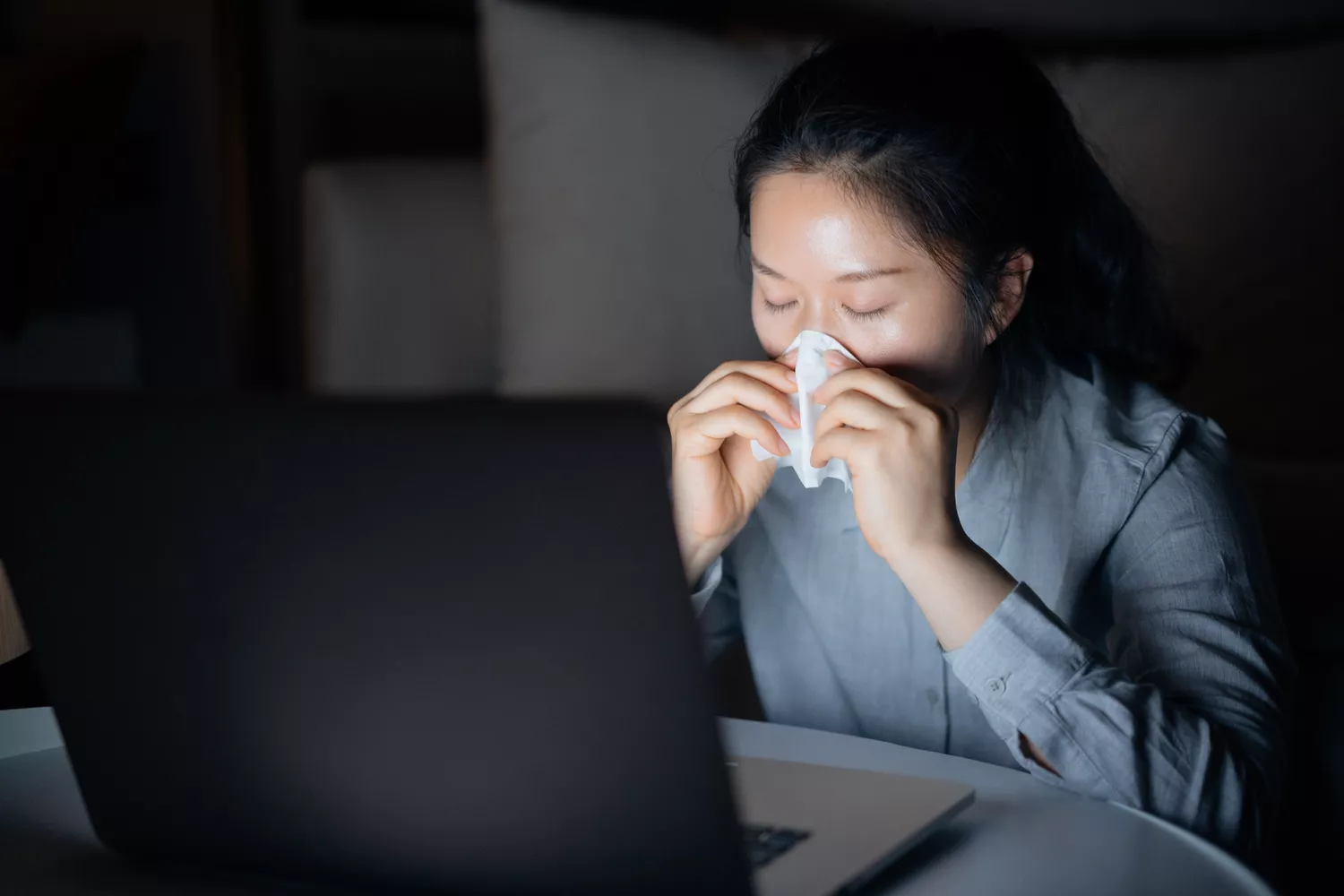 A person in a dark room holding a tissue over their nose and mouth while looking at a laptop screen.