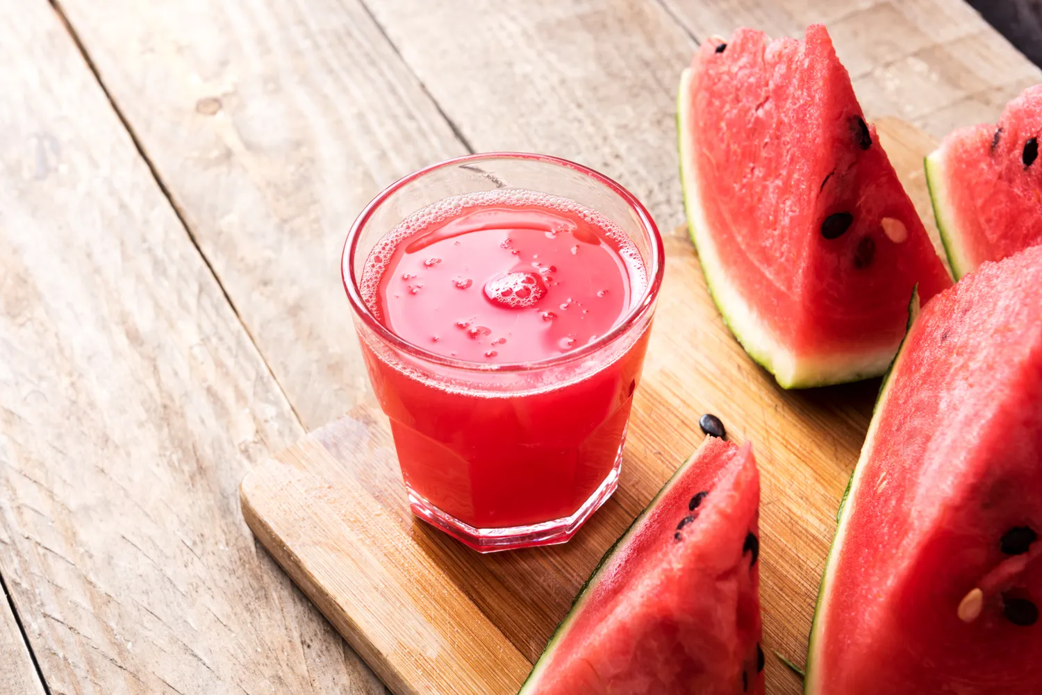 A glass of watermelon juice next to fresh slices of watermelon on a wooden board.