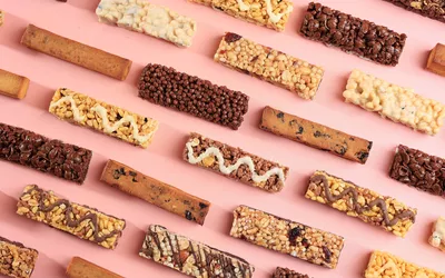 An assortment of various snack bars on a pink background, showcasing processed convenient foods.