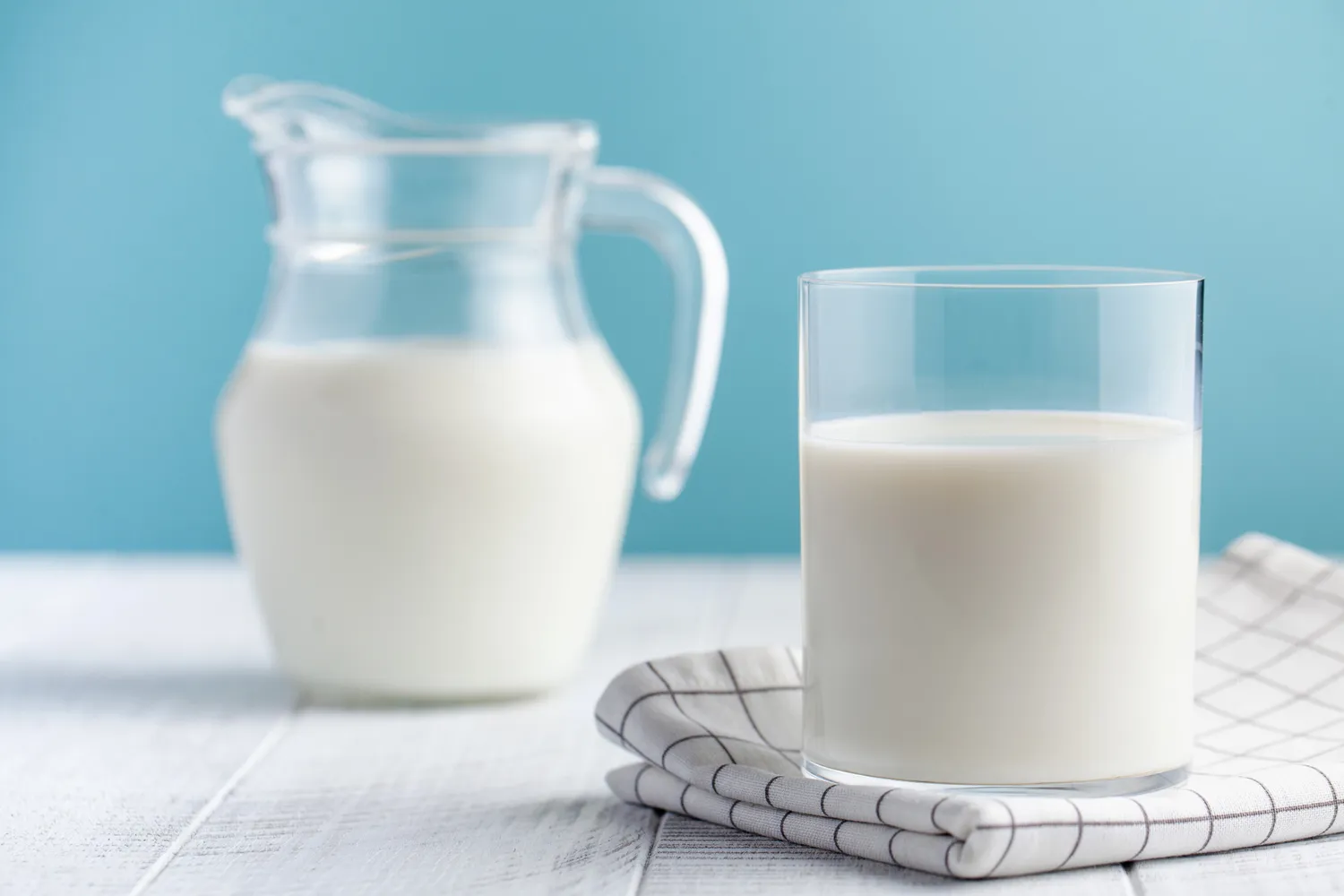 A glass and a jug of fresh milk set against a clean blue background.