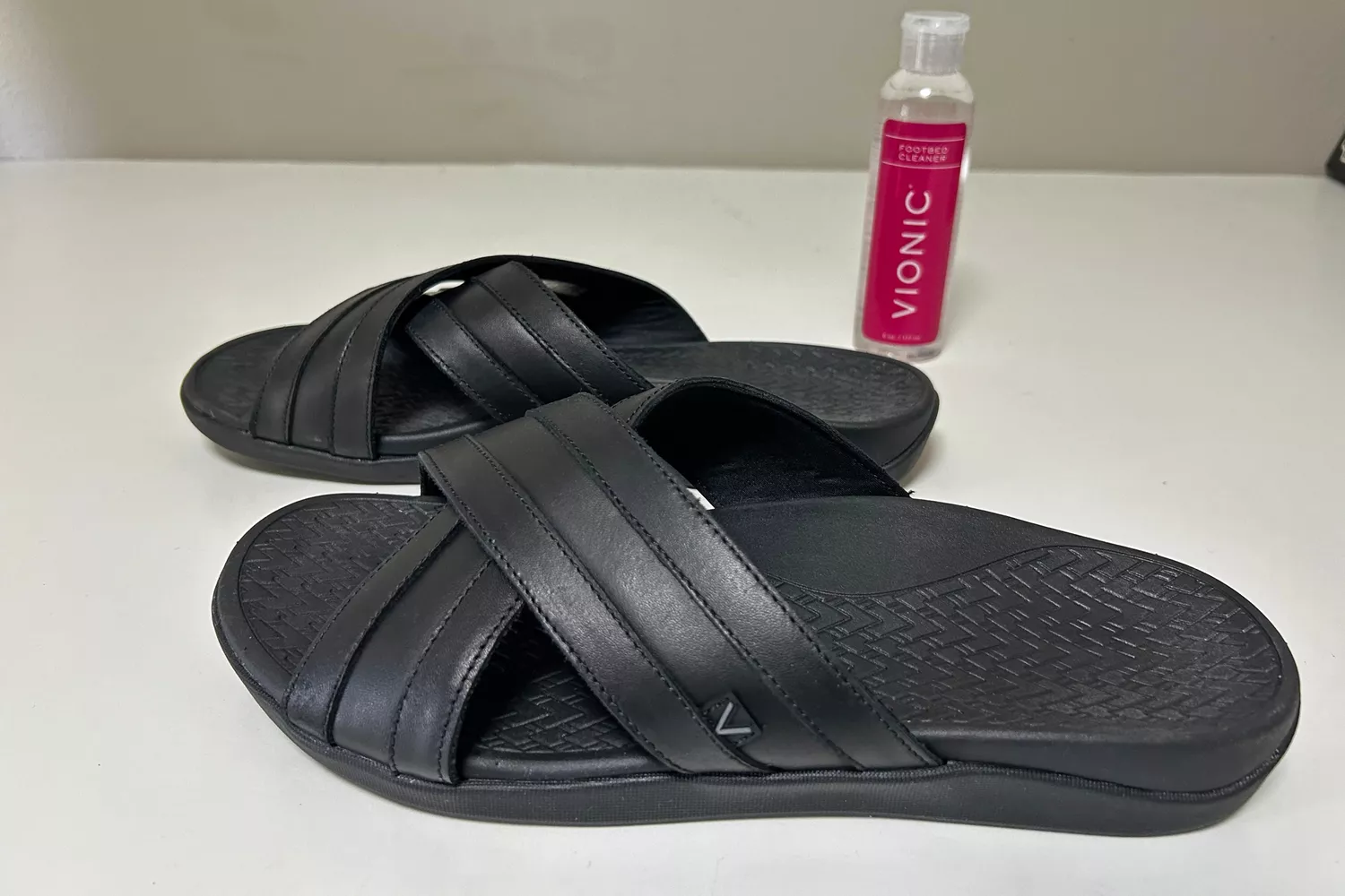 Black Vionic slide sandals placed next to a bottle of Vionic Footbed Cleaner on a clean white surface.
