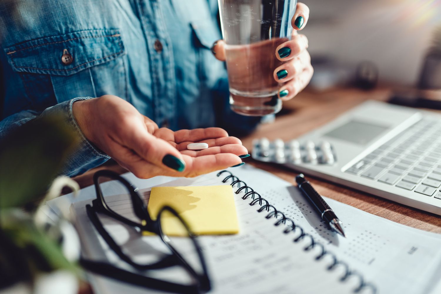 A person with teal nail polish holds a pill and a glass of water at a desk with a laptop and medication pack.