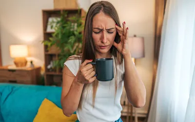 A woman in a living room holding her head in pain, representing a sugar-induced headache or crash.
