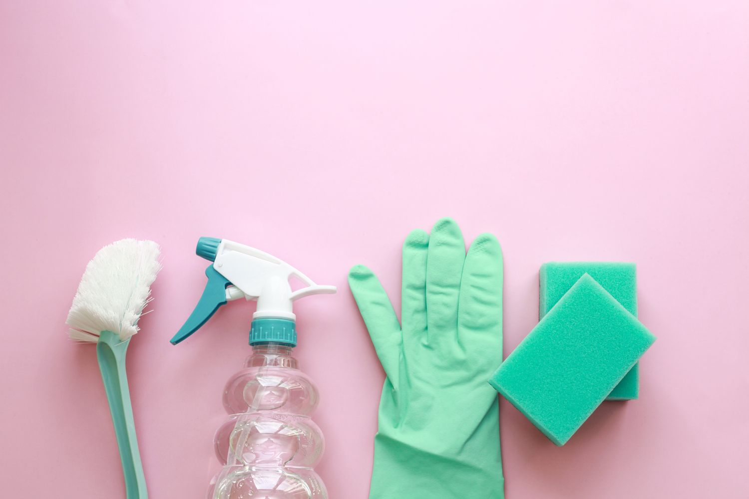 A flat-lay arrangement of cleaning supplies including a spray bottle, brush, and sponges on a pink background.