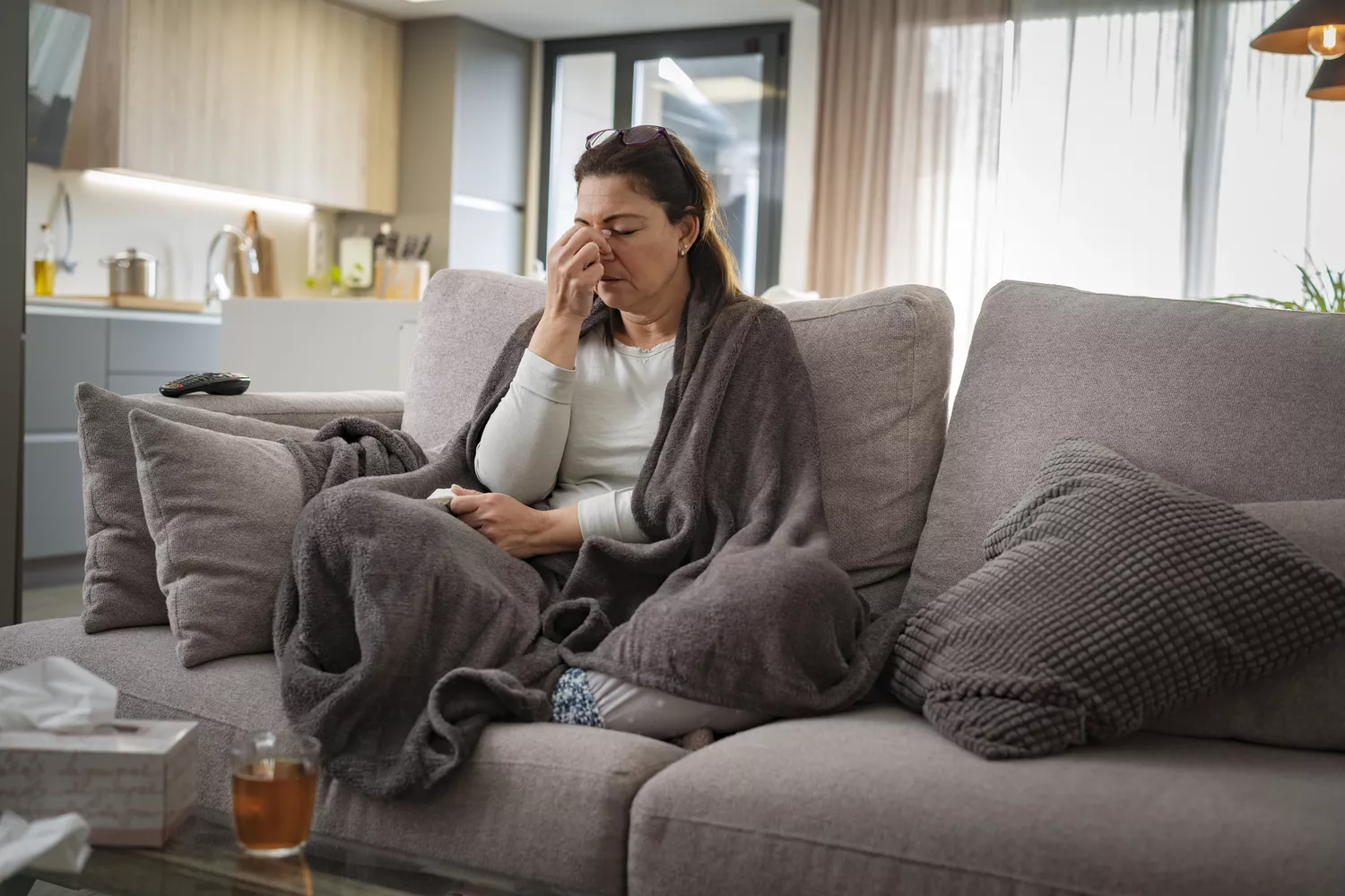 A person wrapped in a blanket on a couch touching the bridge of their nose with a look of discomfort.