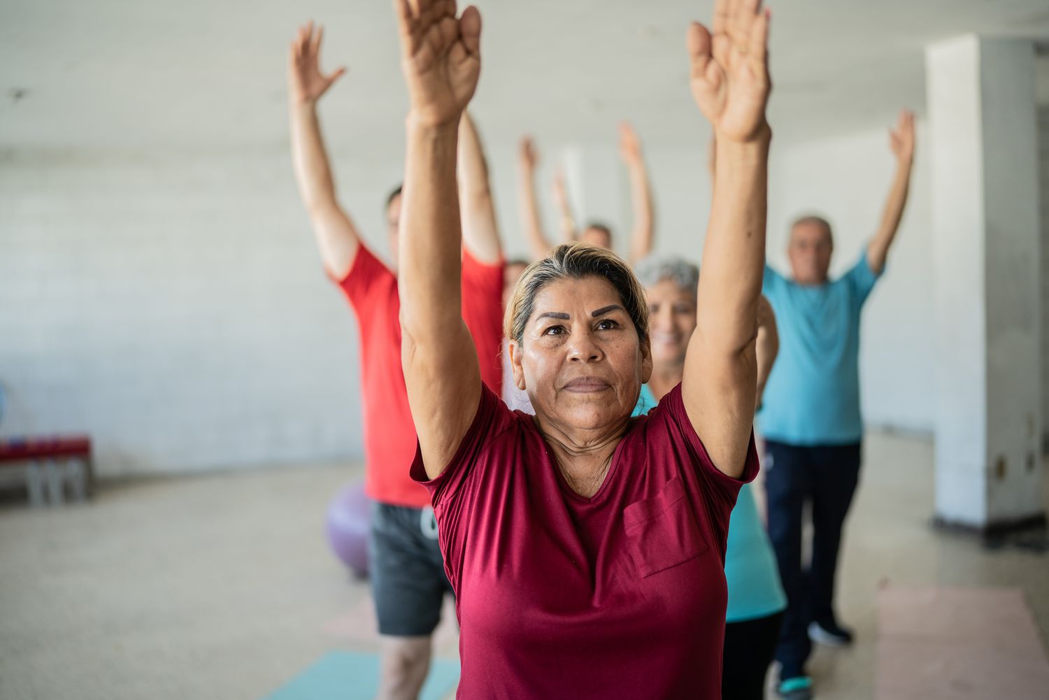 A senior woman performing guided stretching exercises alongside peers in a bright fitness studio.