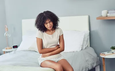 A woman sitting on a bed holding her stomach in discomfort, illustrating digestive pain.