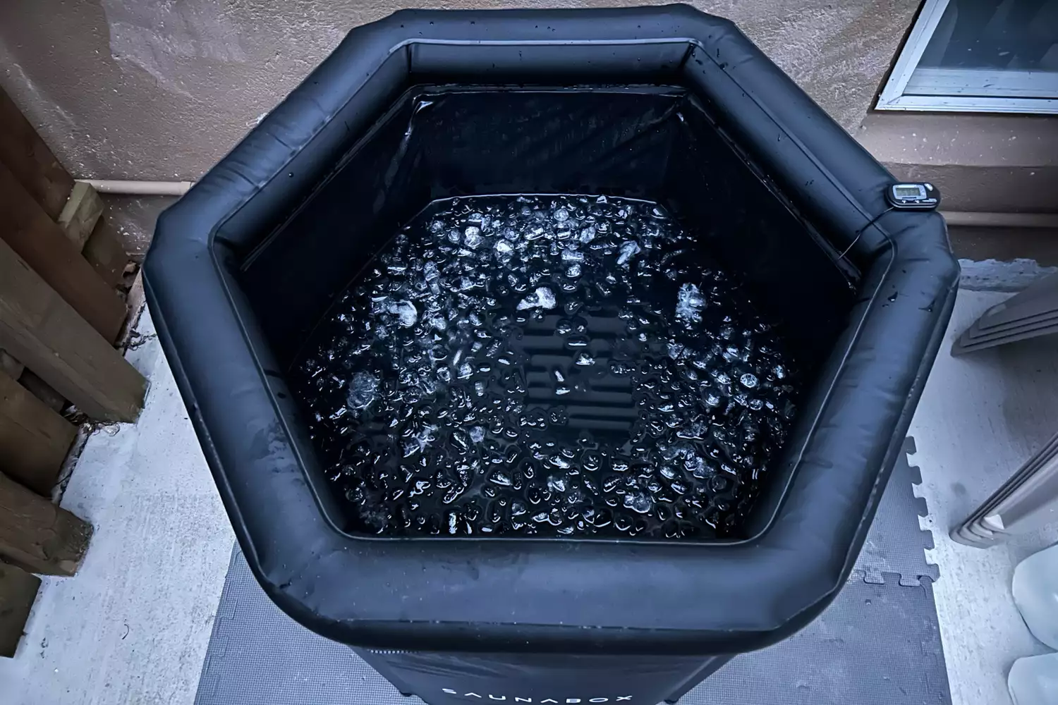 A black hexagonal Saunabox inflatable cold plunge tub filled with ice water on a patio.