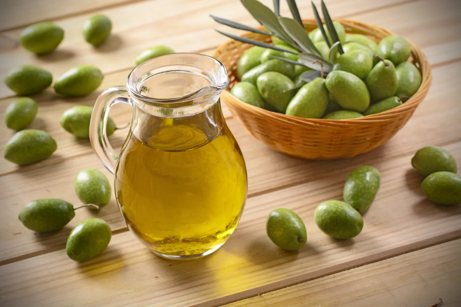 A glass pitcher of olive oil surrounded by fresh green olives.