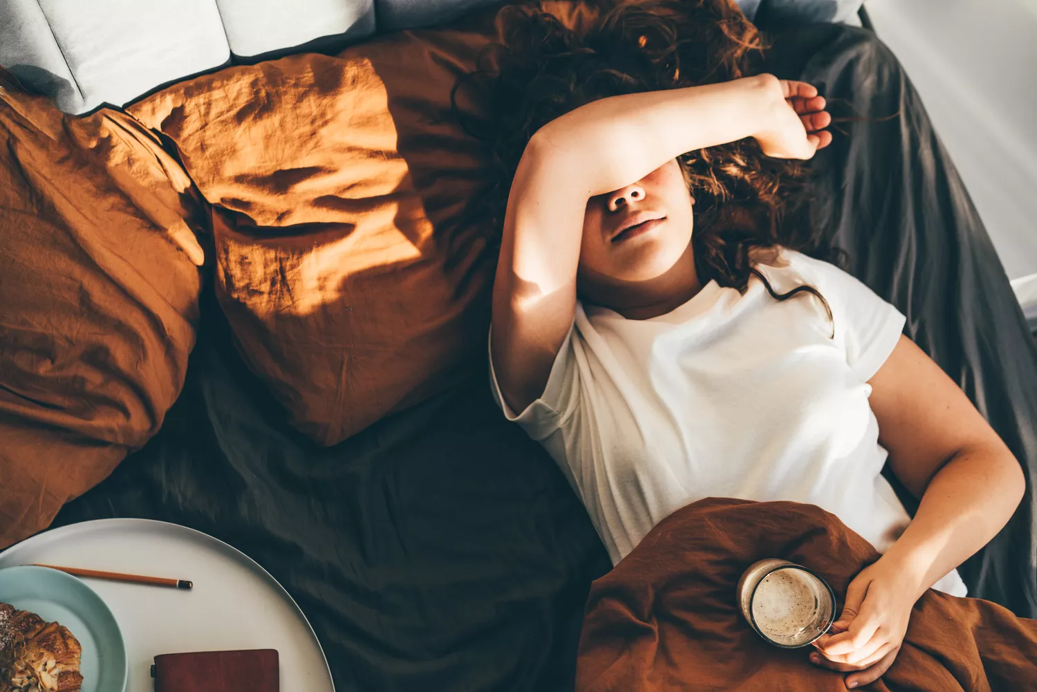 A woman lying in bed with a mug, appearing to rest or recover in a sunlit room.
