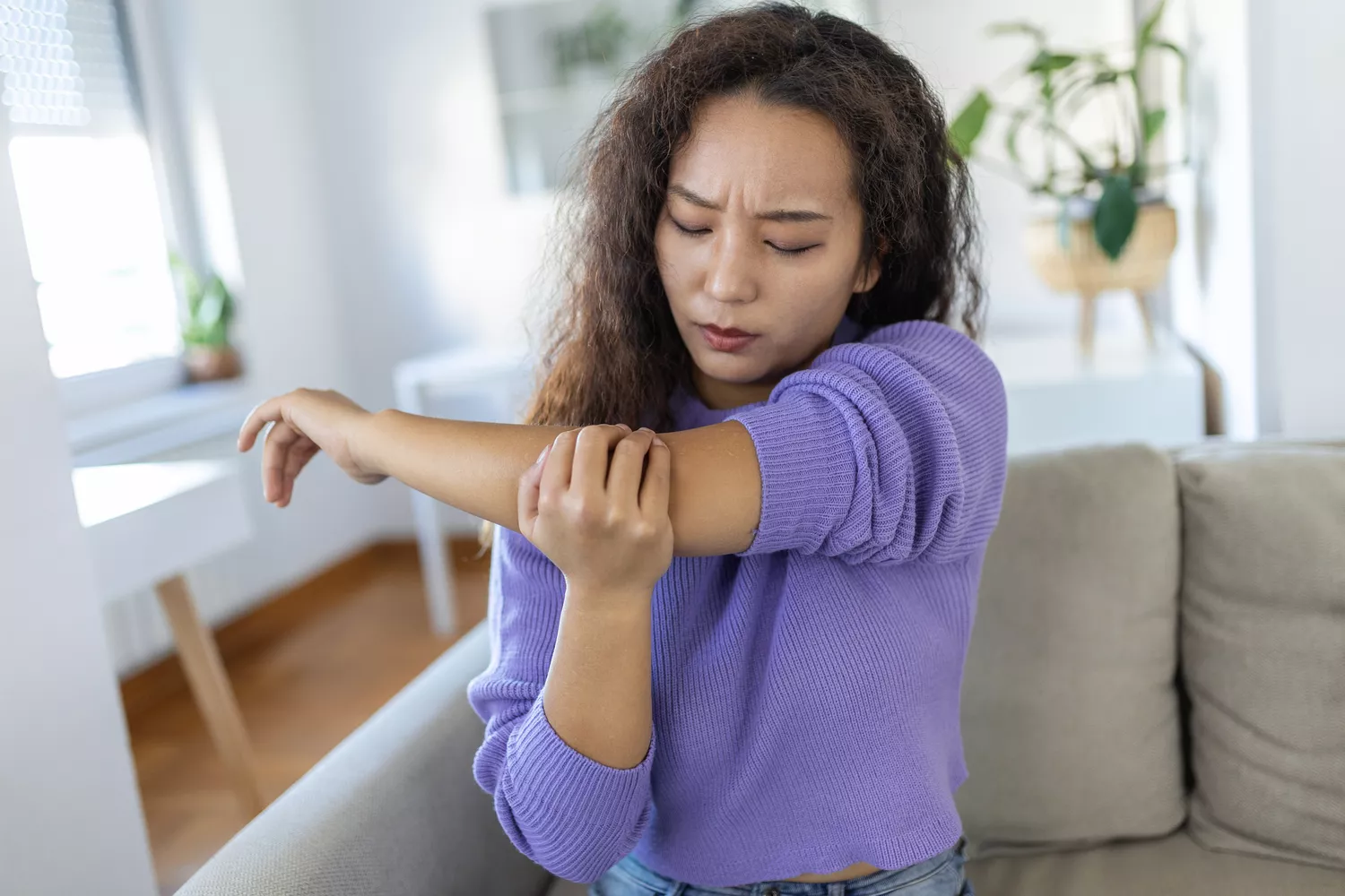 A woman in a purple sweater sitting on a couch, clutching her arm with a grimace of pain.