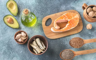 A variety of omega-3 rich foods including salmon, avocado, and walnuts on a grey surface.