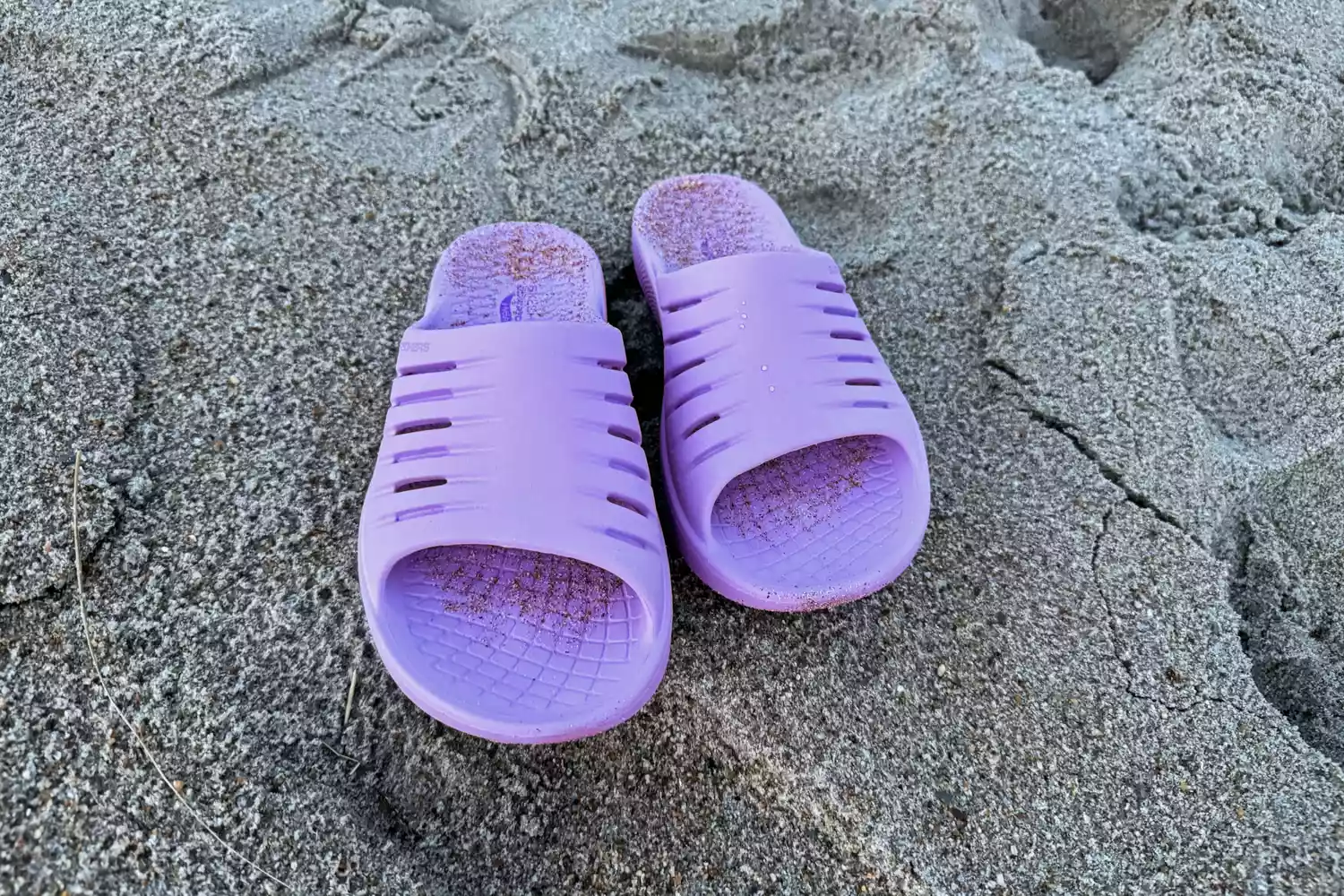 A pair of purple Skechers slides resting on sandy beach terrain near the water.